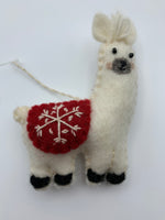 Load image into Gallery viewer, This close-up view of the llama Christmas ornament is hand-crafted (fair trade) and made of 100% natural hand-felted wool. It is off-white with a gray muzzle and black accents (hooves, nose, etc.) has a cute tuft of (yarn) hair between his ears &amp; wears a rounded red blanket on its back with a &#39;signature&#39; (white) snowflake on it. It is approximately 4.5&quot;x3.5&quot;. It comes with a detachable fair trade holiday &quot;to/from&quot; tag to use is giving as a gift.
