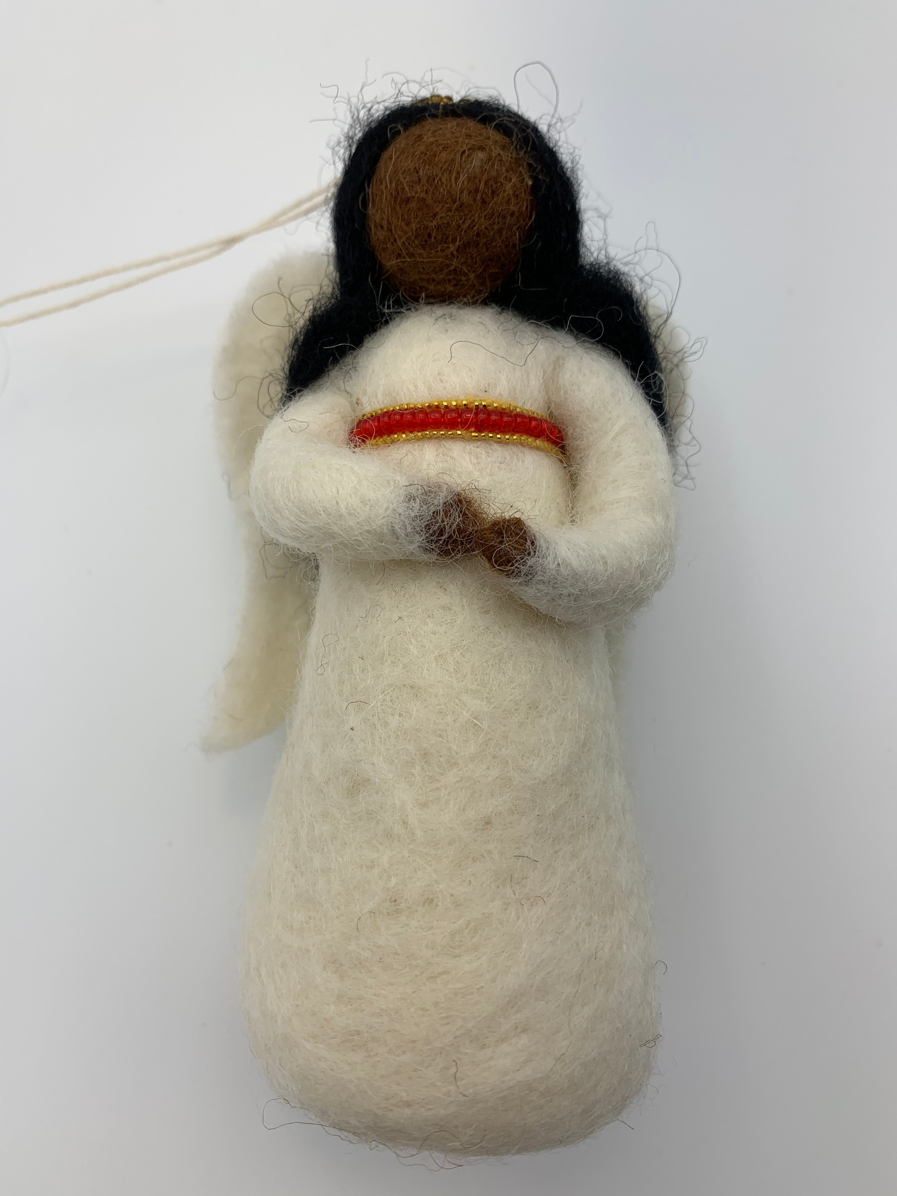 This is an close-up photo of an angel of color (black) Christmas ornament that is handcrafted (fair trade) and made of 100% natural wool. She wears a white dress accented with red and gold beading on empire waist, has a gold beaded halo, black face and hair and bendable arms. Approximately 5"x2.5". She comes with a fair trade holiday "to/from" tag to use if giving this as a gift.