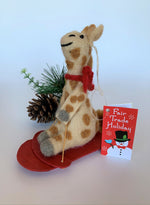 Load image into Gallery viewer, This is a side view of a sledding giraffe Christmas ornament that is handcrafted (fair trade) and made of 100% natural wool. The sled is handmade using paper that is hardened and feels almost like thin wood. It is tan with brown spots, gray accents on feet and hands, with black accents (mouth, eyes, etc.) and wearing a red winter scarf. It sits on a red sled, holding a gold sled &quot;rope.&quot; Approximately 6&quot;x4.75x1.5&quot;. It comes with a fair trade holiday &quot;to/from&quot; tag to use if giving this as a gift.

