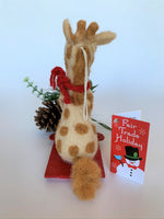Load image into Gallery viewer, This is a back view of a sledding giraffe Christmas ornament that is handcrafted (fair trade) and made of 100% natural wool. The sled is handmade using paper that is hardened and feels almost like thin wood. It is tan with brown spots, gray accents on feet and hands, with black accents (mouth, eyes, etc.) and wearing a red winter scarf. It sits on a red sled, holding a gold sled &quot;rope.&quot; Approximately 6&quot;x4.75x1.5&quot;. It comes with a fair trade holiday &quot;to/from&quot; tag to use if giving this as a gift.
