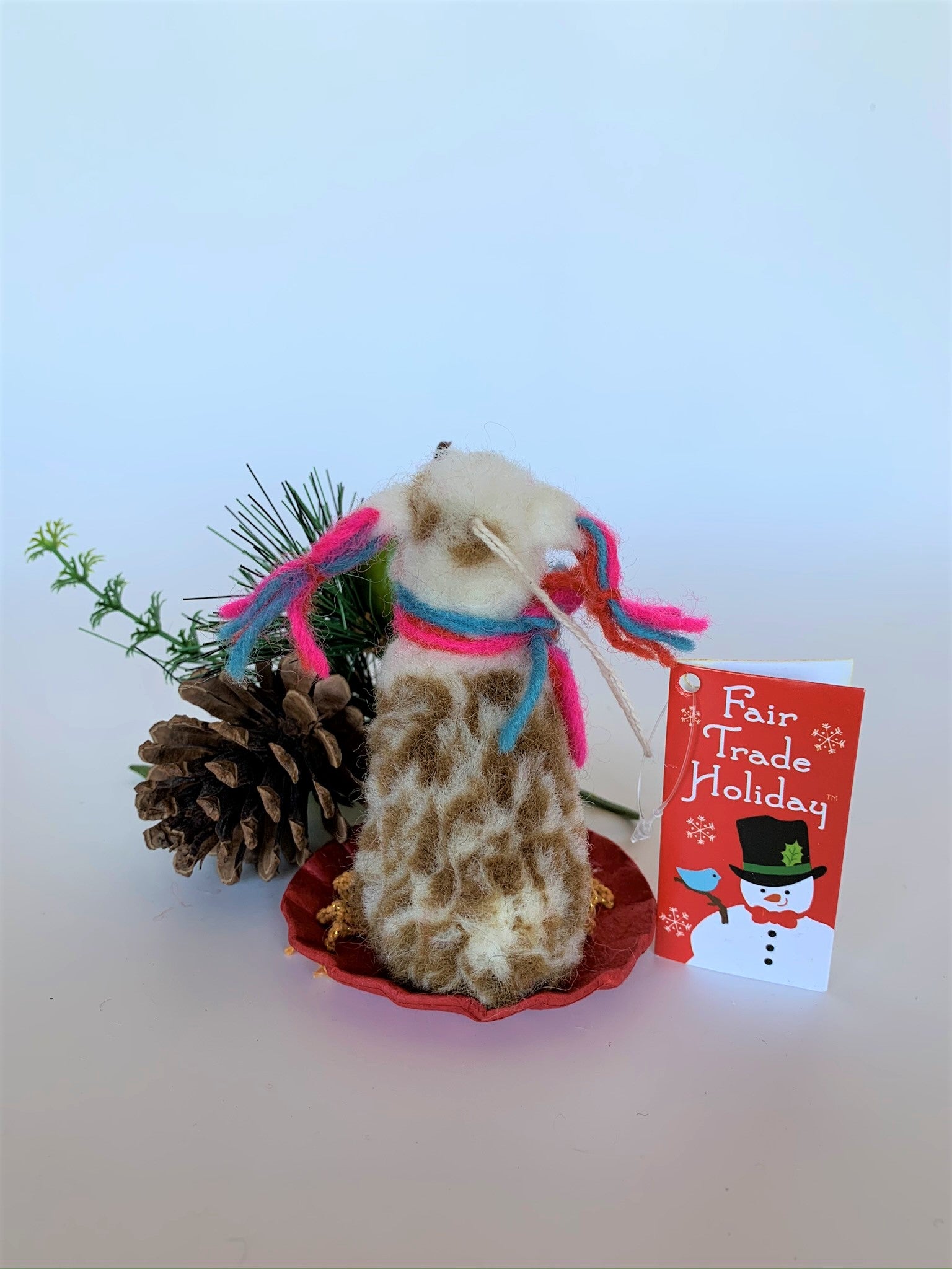 This is a back view of the sledding llama Christmas ornament that is handcrafted (fair trade) and is made of 100% natural, hand-felted wool. The sled is handmade using paper and feels something like very thin wood. The llama is furry, off-white and brown, with black accents (hooves, facial features), sits on a round red sled and wears a scarf made of colorful (red, blue and pink) yarn. The same yarn is used for accents on its ears. Approximately 4.5"x2.75"x2.75".