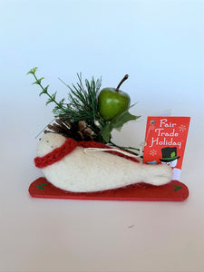 This is a back view of the snowboarding seal Christmas ornament that is handcrafted (fair trade) using 100% natural wool. The snowboard is handmade using paper that is hardened and feels almost like thin wood. It is off-white with black accents (eyes, nose, whiskers etc.), wears a red winter scarf and is lying on a red snowboard with green accents. Approximately 5.5"x2"x1.75". It comes with a fair trade holiday "to/from" tag to use if giving this as a gift.