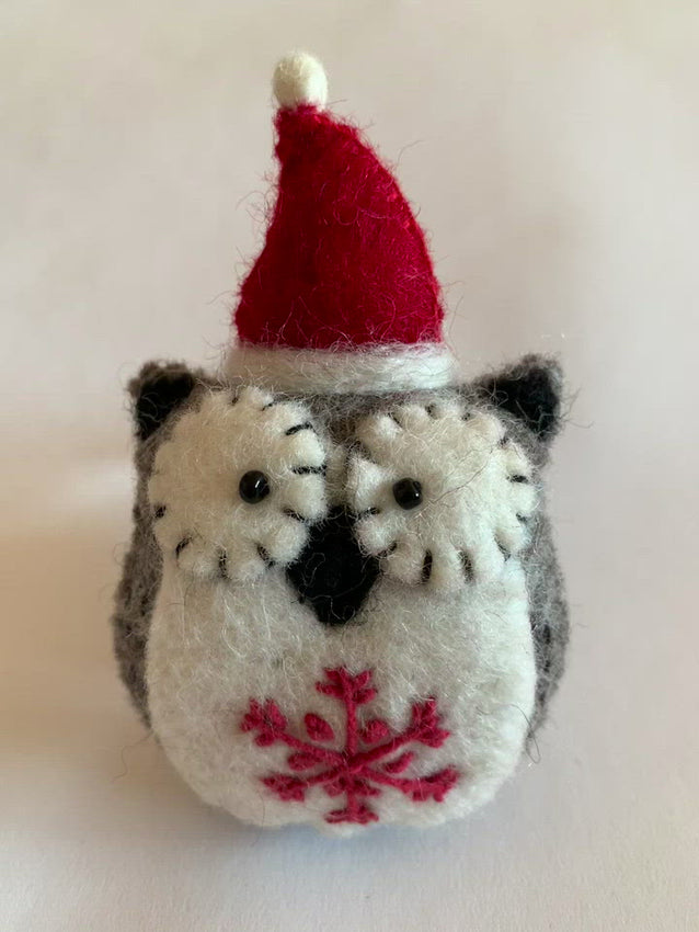 This close-up view of the Santa-hat owl Christmas ornament is handmade (fair trade) from 100% hand-felted wool. It is off-white and brownish-gray with some black accents (beak, ears, etc.) and wears an off-white and red Santa hat. The 'signature' (red) snowflake is displayed on its chest/belly area. It is approximately 4.5"x3" and comes with a detachable fair trade holiday "to/from" tag to use if giving as a gift. 