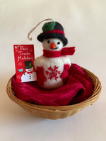 Load and play video in Gallery viewer, This Snowman Christmas ornament is handmade (fair trade) from 100% hand-felted wool. It is off-white  with some black accents (eyes, mouth) and wears a black top-hat with red &amp; green accents and red woolen mittens and scarf. It has a orange carrot shaped nose and the &#39;signature&#39; (red) snowflake is displayed on its belly. It is approximately 4.5&quot;x2.75&quot; and comes with a detachable fair trade holiday &quot;to/from&quot; tag to use if giving as a gift.     
