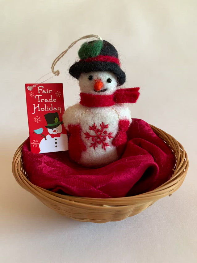 This Snowman Christmas ornament is handmade (fair trade) from 100% hand-felted wool. It is off-white  with some black accents (eyes, mouth) and wears a black top-hat with red & green accents and red woolen mittens and scarf. It has a orange carrot shaped nose and the 'signature' (red) snowflake is displayed on its belly. It is approximately 4.5"x2.75" and comes with a detachable fair trade holiday "to/from" tag to use if giving as a gift.     