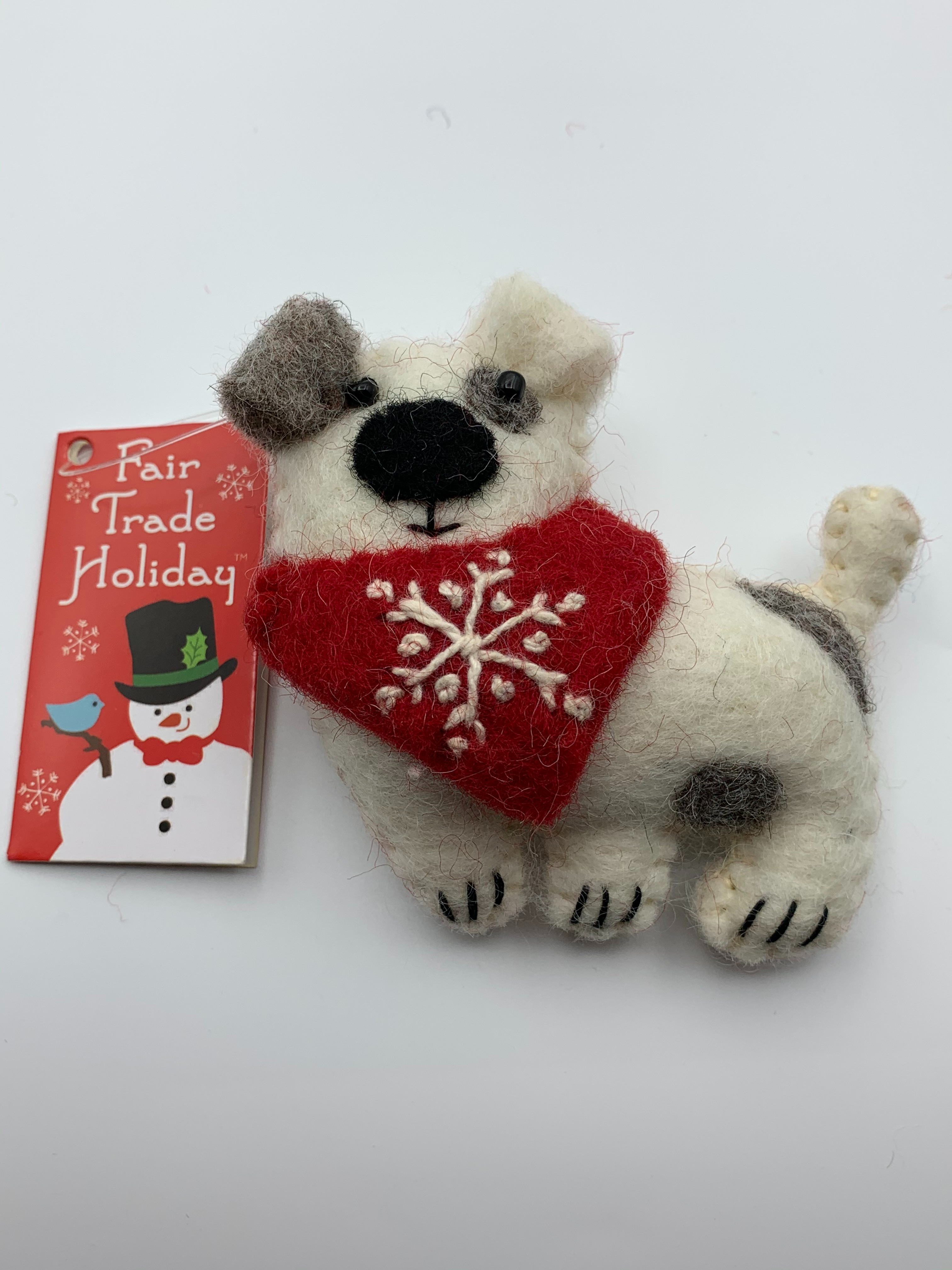 This close-up of Patches the dog is a hand-crafted (fair trade) Christmas ornament made of 100% natural wool. He is off-white with brown patches and a red bandana with a 'signature' (white) snowflake on it. Approximately 3.75"x3.75". He comes with a detachable fair trade holiday tag to use if you are giving as a gift.