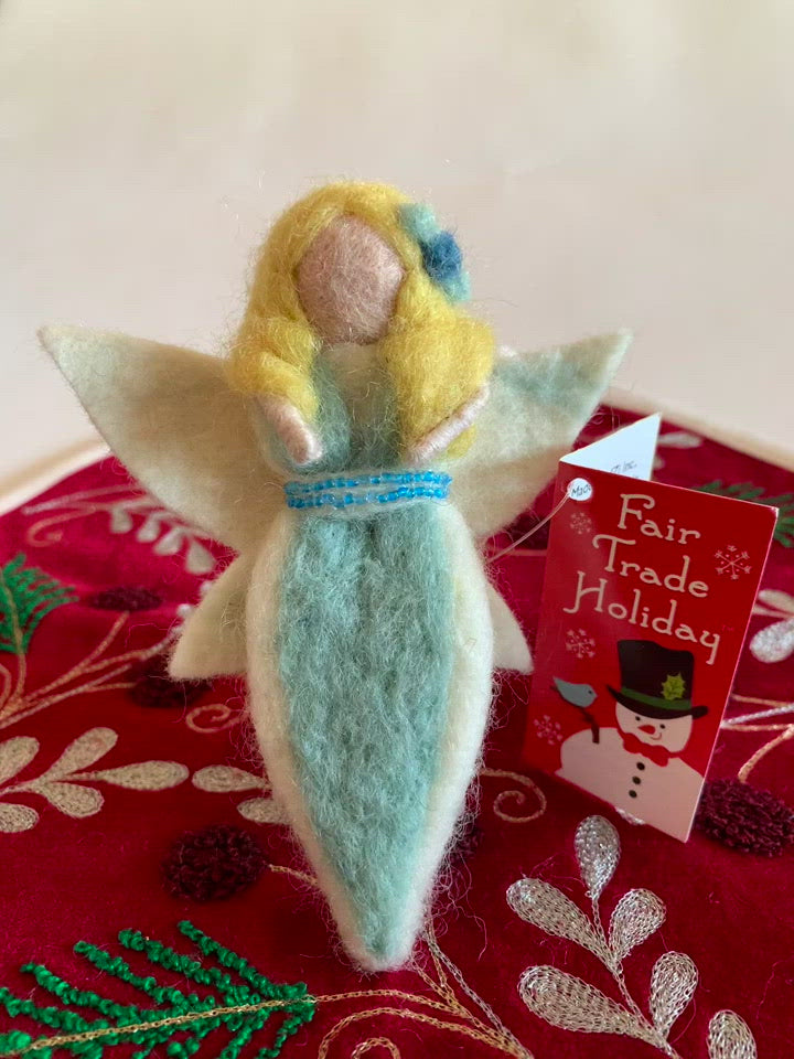 The air-fairy Christmas ornament is handmade (fair trade) from 100% wool. She is light/fair skinned & wearing a light blue dress with white on the edge with a double row of tiny blue beads around the empire waist. She wears a two-toned blue flower in her yellow hair & has white wings with a bit of light yellow on them. She has bendable arms and no facial markings. Approximately 5"x2.5" & comes with a fair trade holiday "to/from" tag to use if giving as a gift. 