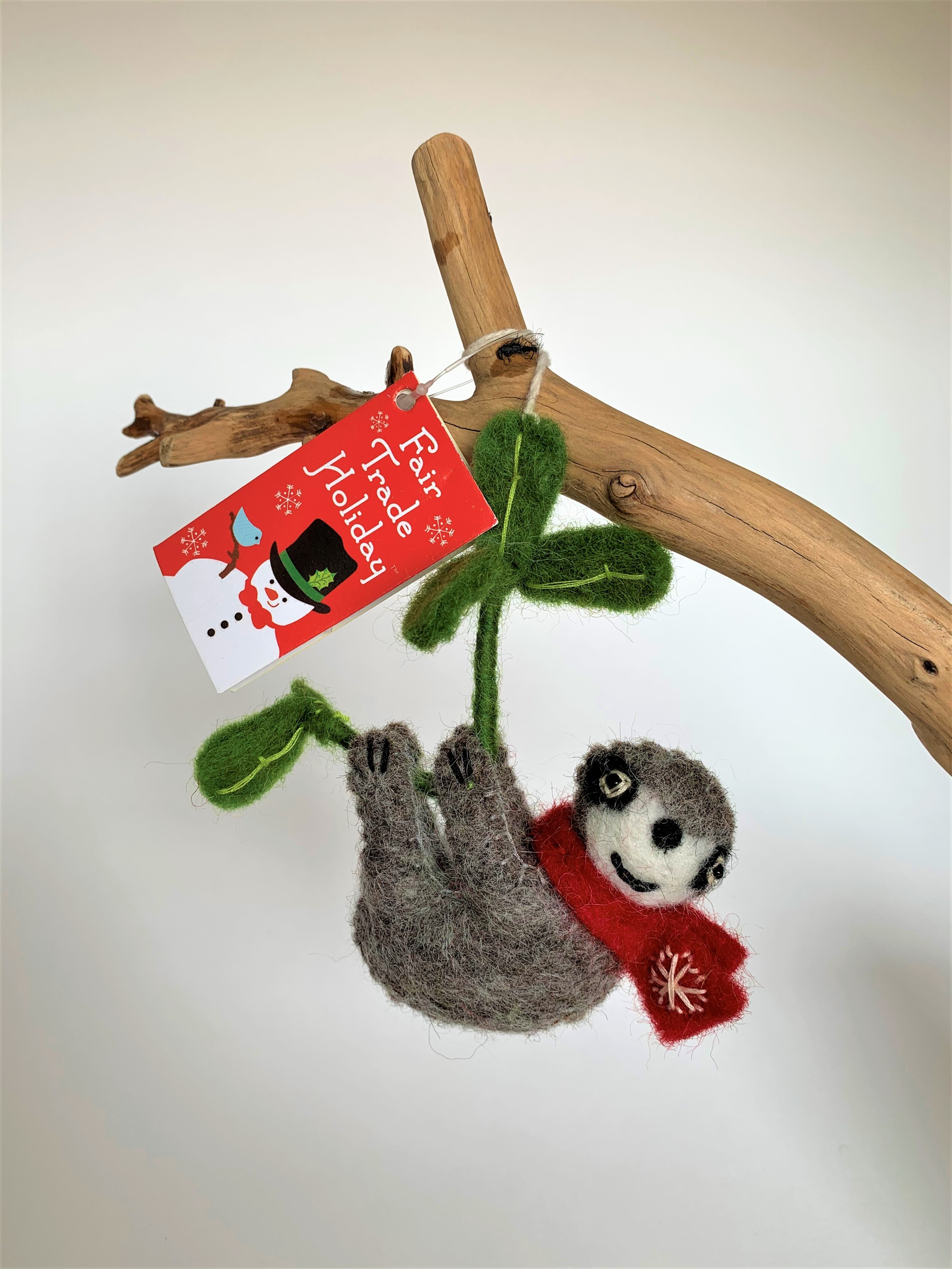 This view of the sloth Christmas ornament, hanging on a tree branch, is hand-crafted (fair trade) and made of 100% natural wool. It is brownish-gray with an off-white face, with black accents (nose, mouth, etc) and is wearing a red winter scarf with the 'signature' (white) snowflake on it. It is hanging from a green branch with green leaves. Approximately 4.75"x4.25". It comes with a detachable fair trade "to/from" tag to use if giving as a gift.
