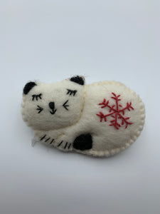 This is a close-up photo of a cat Christmas ornament, curled up and sleeping. It is handcrafted (fair trade) with 100% natural wool. It is off-white with black accents (ears, tail, etc.) and has the 'signature' (red) snowflake on her body. Approximately 3"x4.25" and comes with a detachable fair trade "to/from" tag to use if giving as a gift. 