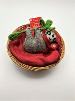 Load image into Gallery viewer, This sloth Christmas ornament is hand-crafted (fair trade) and made of 100% natural wool.  It is brownish-gray with an off-white face, with black accents (nose, mouth, etc) and is wearing a red winter scarf with the &#39;signature&#39; (white) snowflake on it.  It is hanging from a green branch with green leaves. Approximately 4.75&quot;x4.25&quot;. It comes with a detachable fair trade &quot;to/from&quot; tag to use if giving as a gift.
