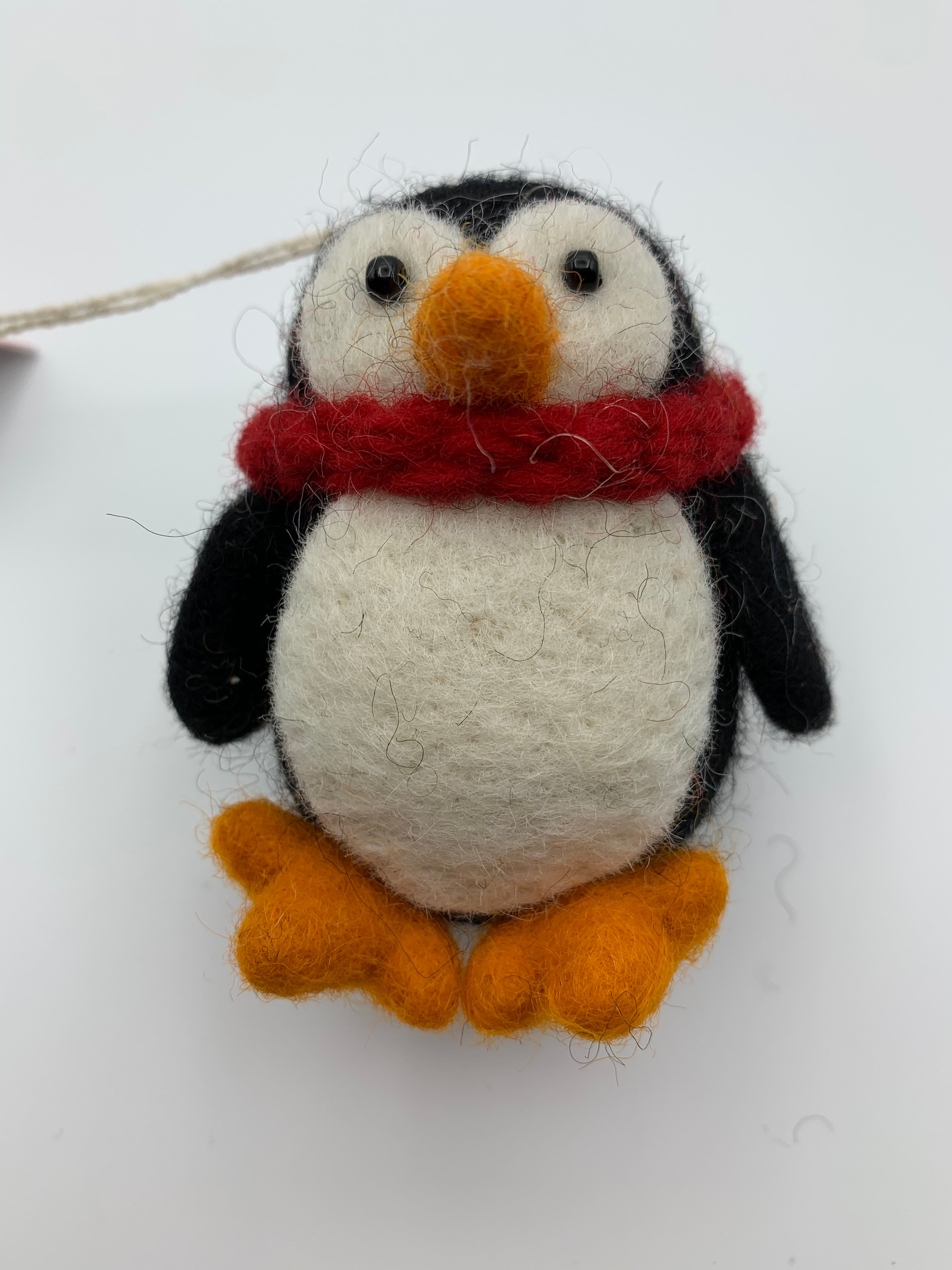 This a close-up view of the Pokey penguin Christmas ornament. It is handcrafted (fair trade), made of 100% hand-felted natural wool, is black and white and a little chubby, with an orange beak and feet, wearing a red winter scarf. Approximately 3.5"x3". He comes with a detachable fair trade holiday tag to use if you are giving this as a gift.