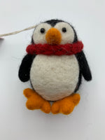 Load image into Gallery viewer, This a close-up view of the Pokey penguin Christmas ornament. It is handcrafted (fair trade), made of 100% hand-felted natural wool, is black and white and a little chubby, with an orange beak and feet, wearing a red winter scarf. Approximately 3.5&quot;x3&quot;. He comes with a detachable fair trade holiday tag to use if you are giving this as a gift.
