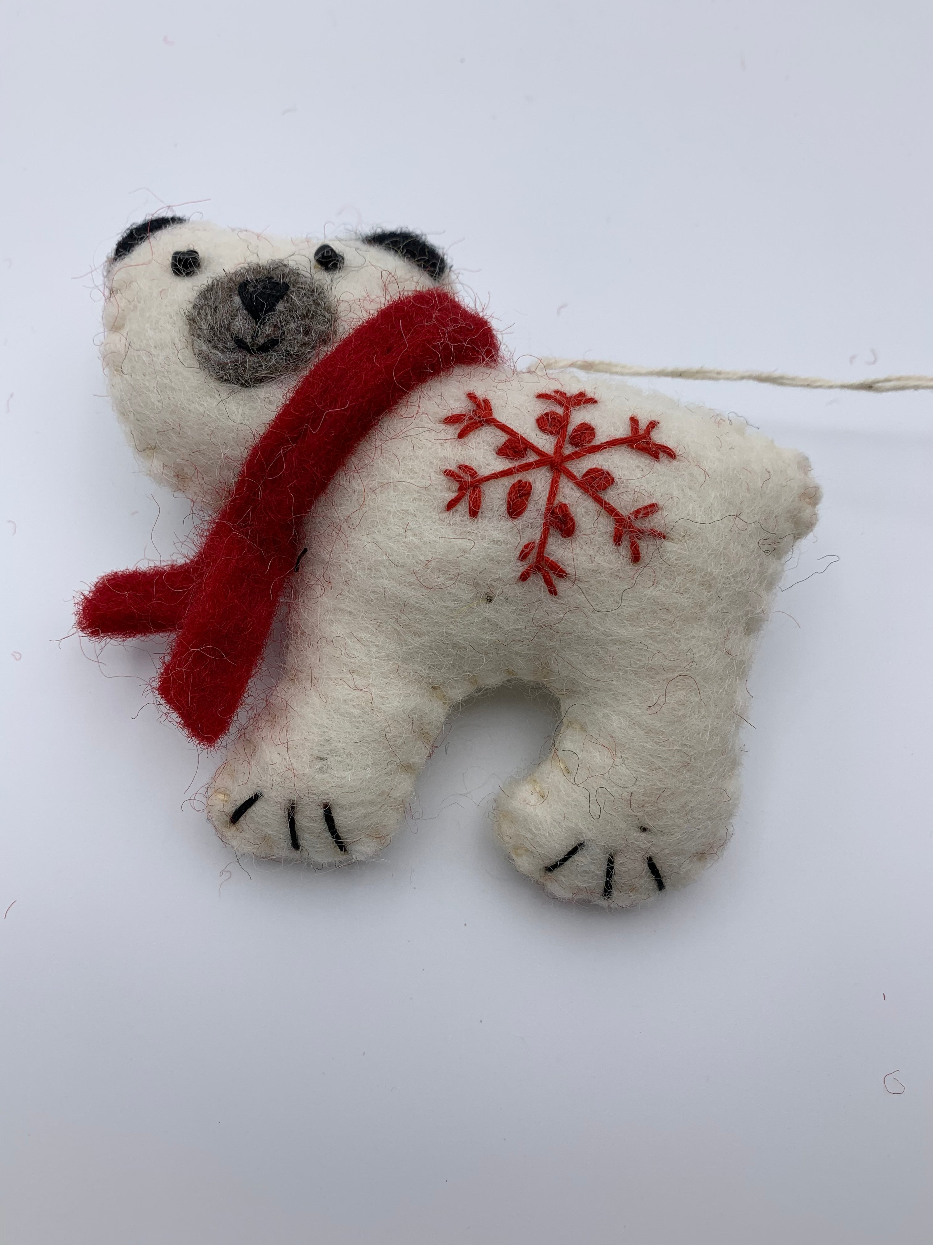 This is a close-up view of the polar bear Christmas ornament that is hand-crafted (fair trade) and made of 100% natural wool. It is off-white with a gray muzzle and black accents (ears, eyes, etc.) and wears a red winter scarf. He has a 'signature' (red) snowflake on his body. Approximately 3.75"x4.2". It comes with a fair trade holiday "to/from" tag to use if giving as a gift.