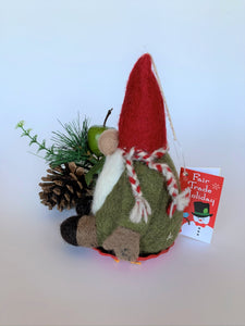  This is a side view of the sledding gnome Christmas ornament that is handcrafted (fair trade) and made of 100% natural wood. The sled is handmade using paper that is hardened and feels almost like thin wood. He wears a green top, brownish-gray pants, a pointed red hat, which covers his eyes, a round tan nose and sits on a round, red sled that has gold colored sled handles on either side. Approximately 6.5"x3.75"x3.5". He comes with a fair trade holiday "to/from" tag to use if giving this as a gift.