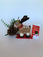 Load image into Gallery viewer, This is a side view of the skiing Rudolph the red-nosed reindeer Christmas ornament that is handcrafted (fair trade) from 100% natural wool. He is off-white and brown with brown ears and tail, dark brown antlers, black accents (feet, mouth, etc.) and a red nose. He wears a red and green winter scarf with an accent of holly and stands on a pair of red skis with green accents on the front. Approximately 3.5&quot;x1.5&quot;x1.75&quot;. He comes with a detachable fair trade holiday &quot;to/from&quot; tag to use if giving as a gift.
