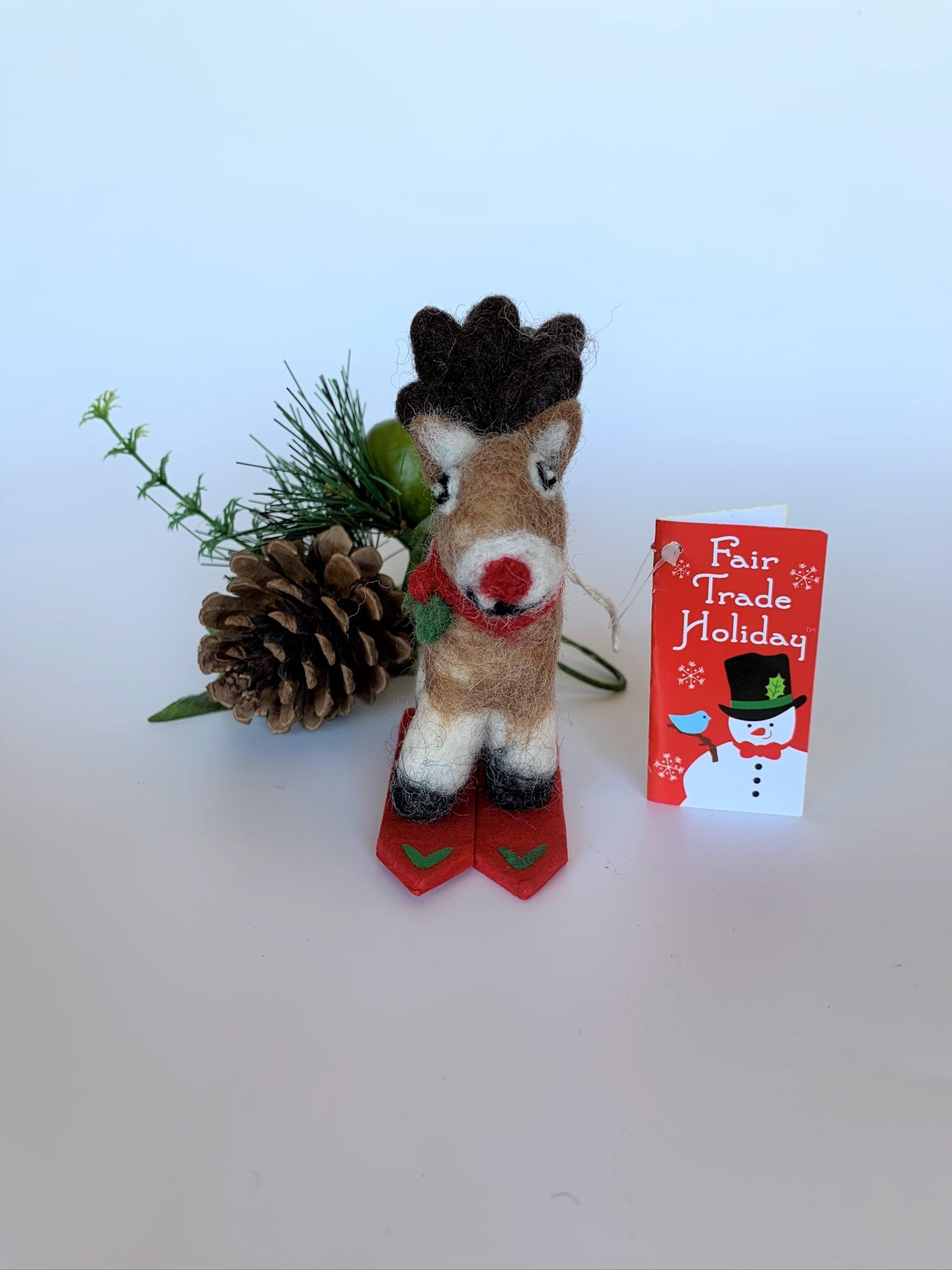 This is a skiing Rudolph the red-nosed reindeer Christmas ornament that is handcrafted (fair trade) from 100% natural wool. He is  off-white and brown with brown ears and tail, dark brown antlers, black accents (feet, mouth, etc.) and a red nose. He wears a red and green winter scarf with an accent of holly and  stands on a pair of red skis with green accents on the front. Approximately 3.5"x1.5"x1.75".  He comes with a detachable fair trade holiday "to/from" tag to use if giving as a gift.