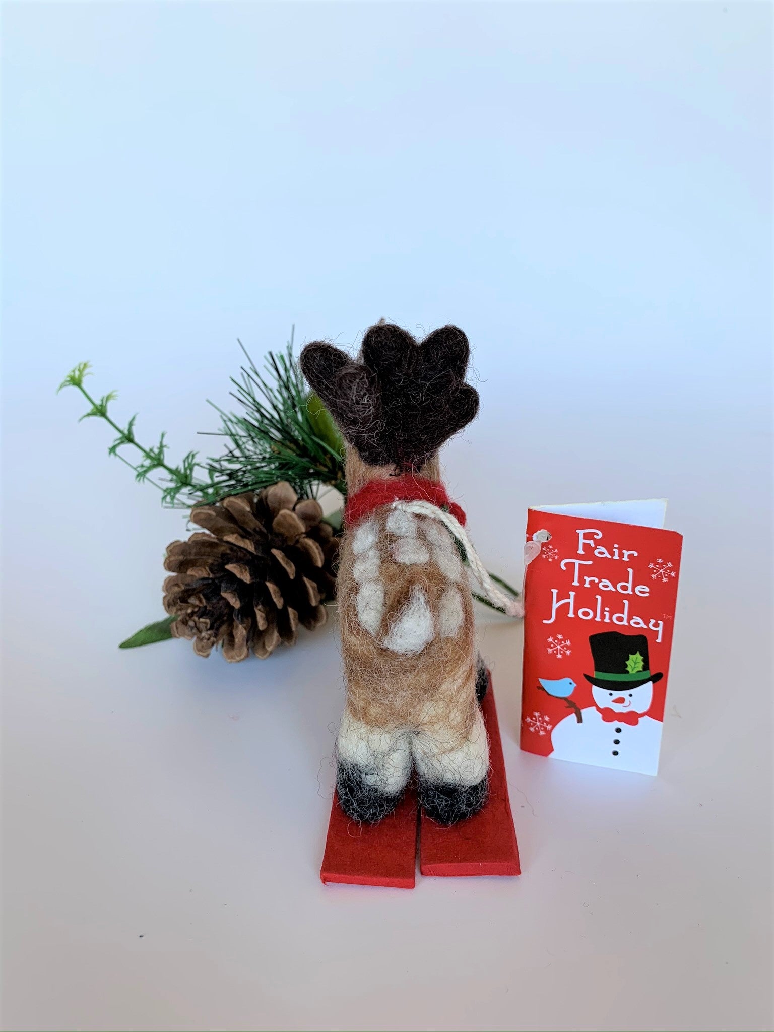This is a back view of the skiing Rudolph the red-nosed reindeer Christmas ornament that is handcrafted (fair trade) from 100% natural wool. He is off-white and brown with brown ears and tail, dark brown antlers, black accents (feet, mouth, etc.) and a red nose. He wears a red and green winter scarf with an accent of holly and stands on a pair of red skis with green accents on the front. Approximately 3.5"x1.5"x1.75". He comes with a detachable fair trade holiday "to/from" tag to use if giving as a gift.