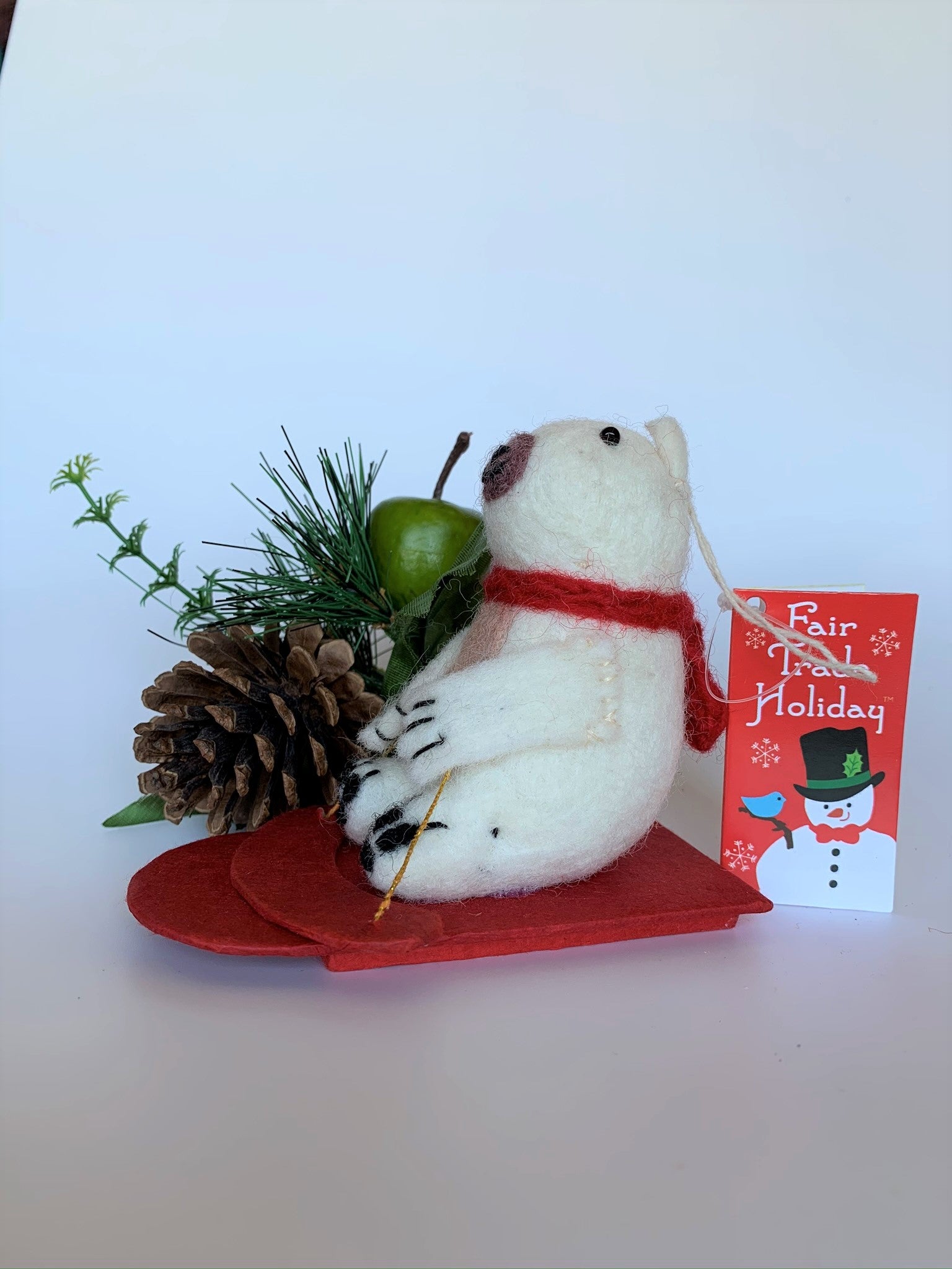 This is a side view of the sledding polar bear Christmas Ornament that is handcrafted (fair trade). The polar bear is made of 100% natural wool and the sled is handmade with paper that is hardened and feels somewhat like very thin wood and it is red. The bear is off-white with a tan belly, brownish-gray muzzle, black accents (paws, nose, etc.) and is wearing a red winter scarf. It is approximately 4.75"x3.5"x2.5" It comes with a fair trade holiday "to/from" tag to use if giving this as a gift.