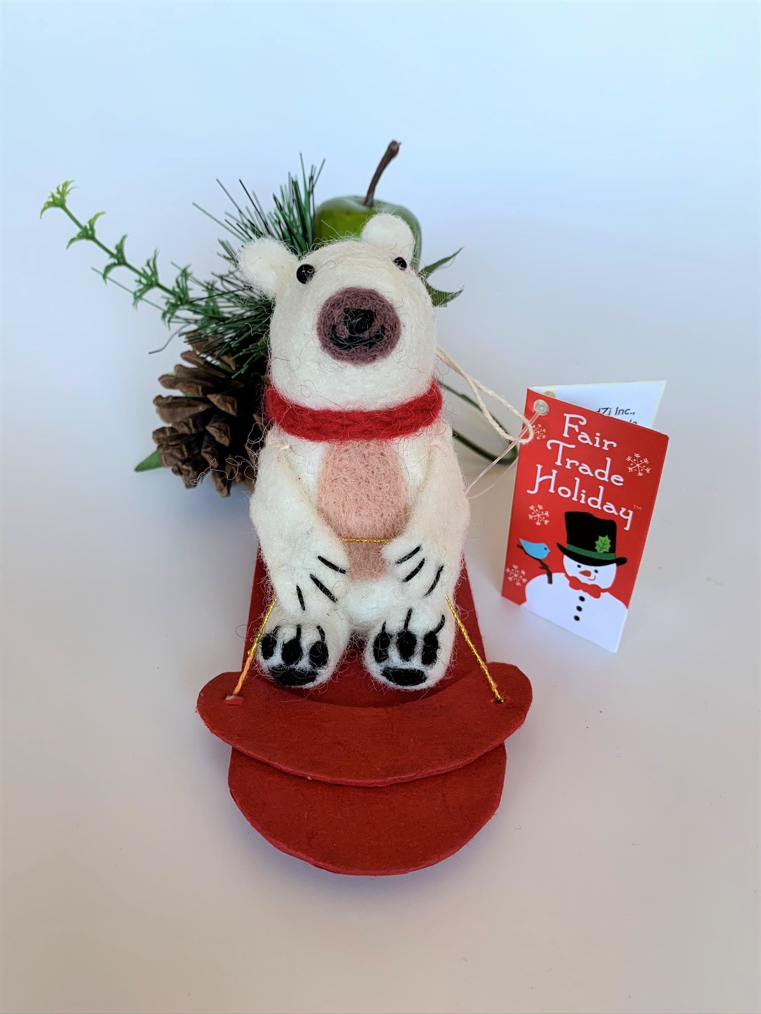 This sledding polar bear Christmas Ornament is handcrafted (fair trade). The polar bear is made of 100% natural wool and the sled is handmade with paper that is hardened and feels somewhat like very thin wood and it is red.  The bear is off-white with a tan belly, brownish-gray muzzle, black accents (paws, nose, etc.) and is wearing a red winter scarf.  It is approximately 4.75"x3.5"x2.5". It comes with a fair trade holiday "to/from" tag to use if giving this as a gift.