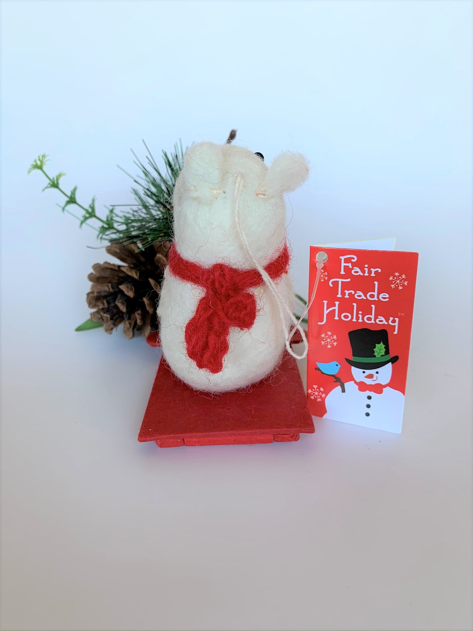 This is a back view of the sledding polar bear Christmas Ornament that is handcrafted (fair trade). The polar bear is made of 100% natural wool and the sled is handmade with paper that is hardened and feels somewhat like very thin wood and it is red. The bear is off-white with a tan belly, brownish-gray muzzle, black accents (paws, nose, etc.) and is wearing a red winter scarf. It is approximately 4.75"x3.5"x2.5". It comes with a fair trade holiday "to/from" tag to use if giving this as a gift.