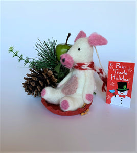 This is a side view of Piggles the sledding piggy Christmas ornament who is handcrafted (fair trade) and made of 100% natural wool. It sits on a round, red sled that is handmade using paper which is hardened and feels almost like thin wood. Piggles is off-white with pink ears, snout and with other pink and black accents (eyes, mouth, etc.). and wears a red & white winter scarf. Approximately 4"x2.75"x2.5". She comes with a detachable fair trade holiday "to/from" tag to use if giving this as a gift.