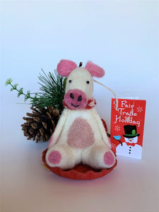 This is Piggles the sledding piggy Christmas ornament who is handcrafted (fair trade) and made of 100% natural wool.  It sits on a round, red sled that is handmade using paper which is hardened and feels almost like thin wood.  Piggles is off-white with pink ears, snout and with other pink and black accents (eyes, mouth, etc.). and wears a red & white winter scarf. Approximately 4"x2.75"x2.5".  She comes with a detachable fair trade holiday "to/from" tag to use if giving this as a gift.