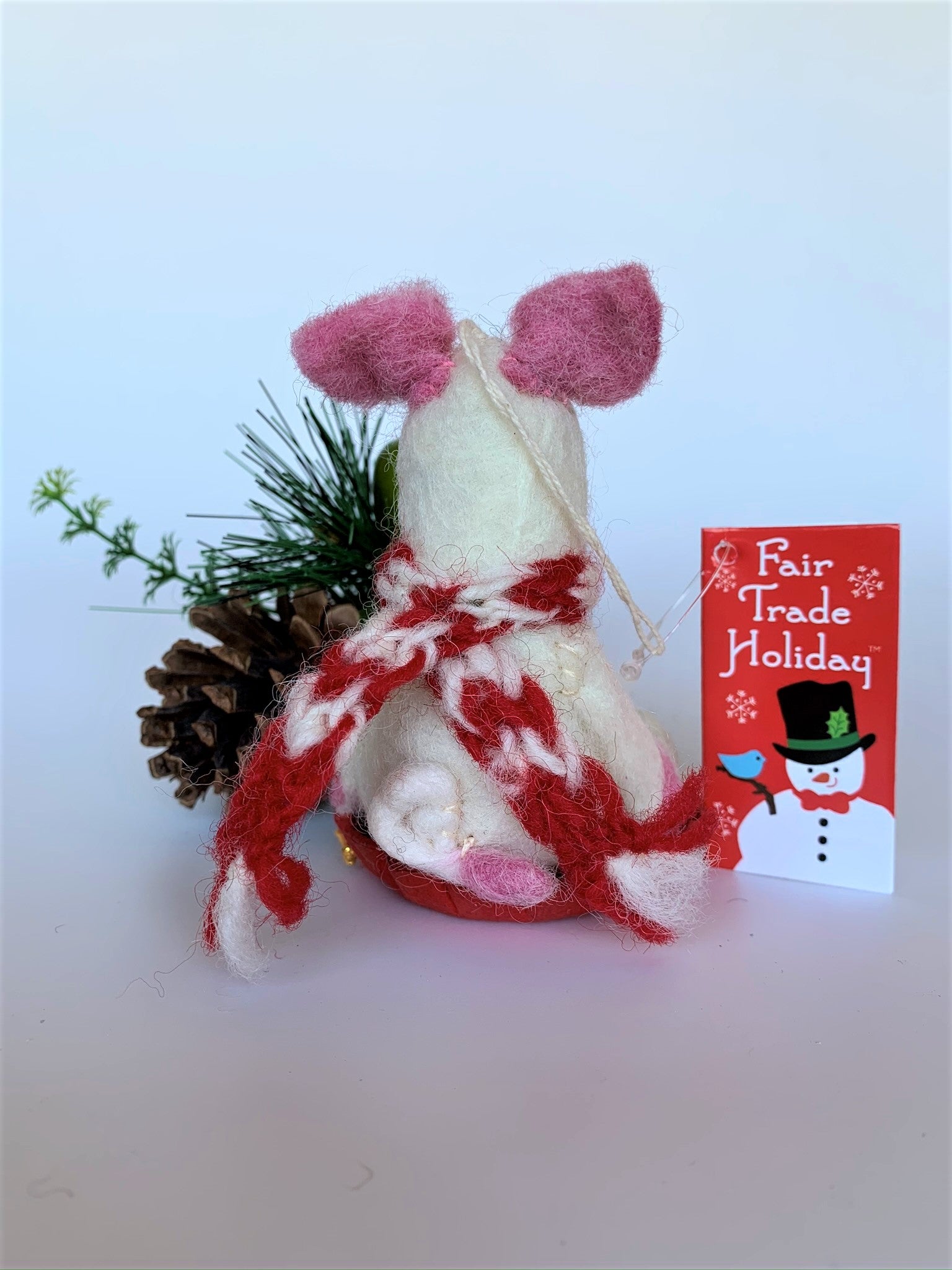 This is a back view of Piggles the sledding piggy Christmas ornament who is handcrafted (fair trade) and made of 100% natural wool. It sits on a round, red sled that is handmade using paper which is hardened and feels almost like thin wood. Piggles is off-white with pink ears, snout and with other pink and black accents (eyes, mouth, etc.). and wears a red & white winter scarf. Approximately 4"x2.75"x2.5". She comes with a detachable fair trade holiday "to/from" tag to use if giving this as a gift.