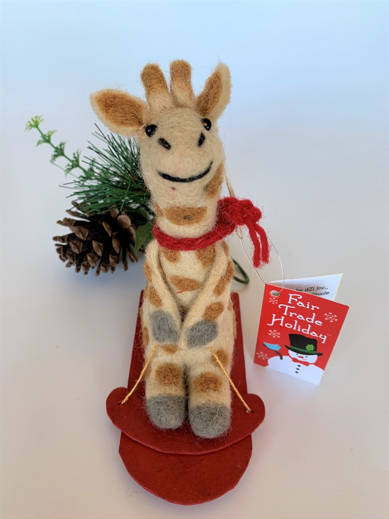 This is a sledding giraffe Christmas ornament that is handcrafted (fair trade) and made of 100% natural wool.  The sled is handmade using paper that is hardened and feels almost like thin wood.  It is tan with brown spots, gray accents on feet and hands, with black accents (mouth, eyes, etc.) and wearing a red winter scarf.  It sits on a red sled, holding a gold sled "rope."  Approximately 6"x4.75x1.5". It comes with a fair trade holiday "to/from" tag to use if giving this as a gift.