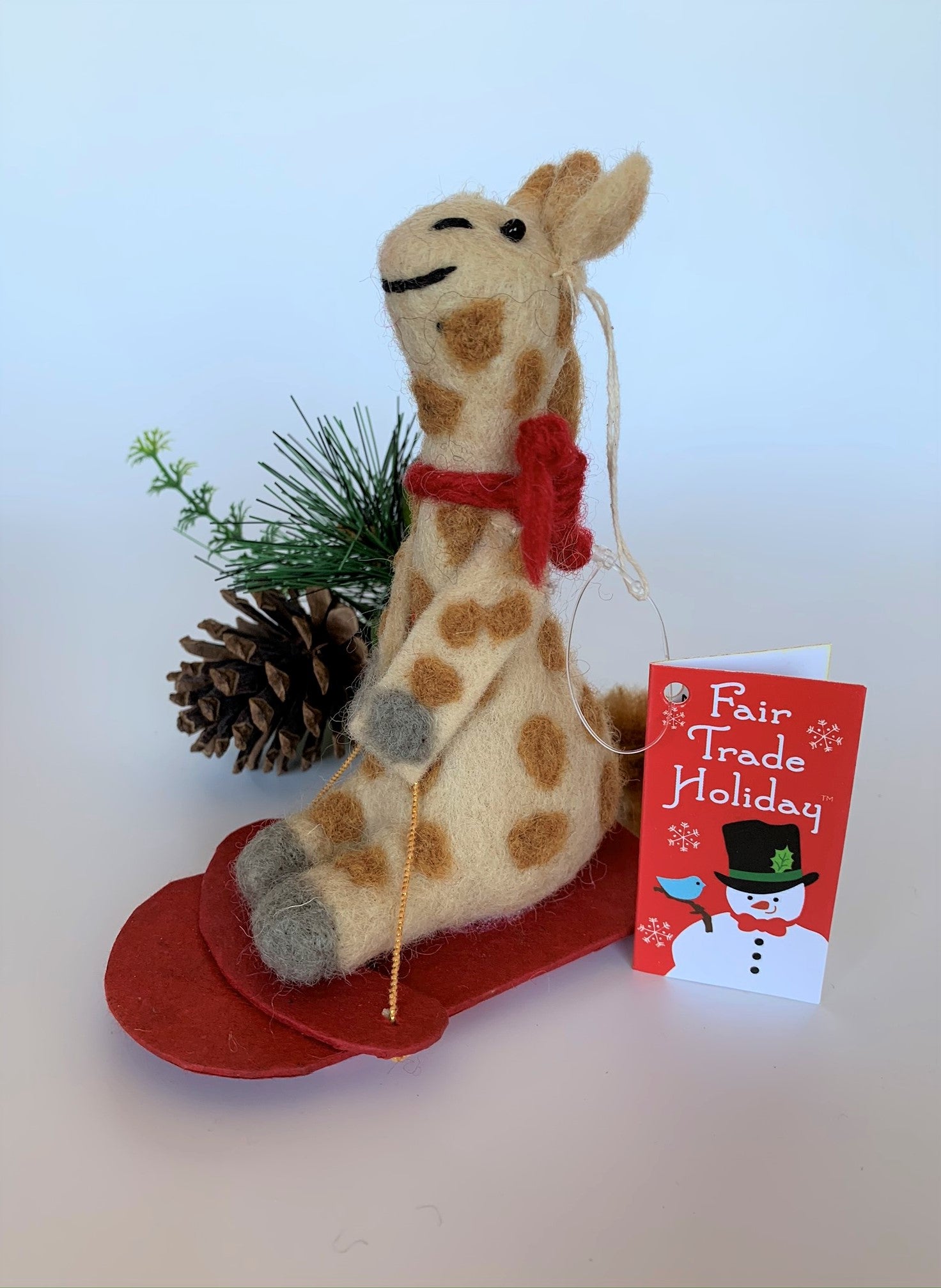 This is a side view of a sledding giraffe Christmas ornament that is handcrafted (fair trade) and made of 100% natural wool. The sled is handmade using paper that is hardened and feels almost like thin wood. It is tan with brown spots, gray accents on feet and hands, with black accents (mouth, eyes, etc.) and wearing a red winter scarf. It sits on a red sled, holding a gold sled "rope." Approximately 6"x4.75x1.5". It comes with a fair trade holiday "to/from" tag to use if giving this as a gift.