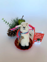 Load image into Gallery viewer, This sledding llama Christmas ornament is handcrafted (fair trade) and is made of 100% natural, hand-felted wool.  The sled is handmade using paper and feels something like very thin wood. The llama is furry, off-white and brown, with black accents (hooves, facial features), sits on a round red sled and wears a scarf made of colorful (red, blue and pink) yarn.  The same yarn is used for accents on its ears.  Approximately 4.5&quot;x2.75&quot;x2.75&quot;.
