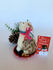  This is a side view of the sledding llama Christmas ornament that is handcrafted (fair trade) and is made of 100% hand-felted  natural wool. The sled is handmade using paper and feels something like very thin wood. The llama is furry, off-white and brown, with black accents (hooves, facial features), sits on a round red sled and wears a scarf made of colorful (red, blue and pink) yarn. The same yarn is used for accents on its ears. Approximately 4.5"x2.75"x2.75".