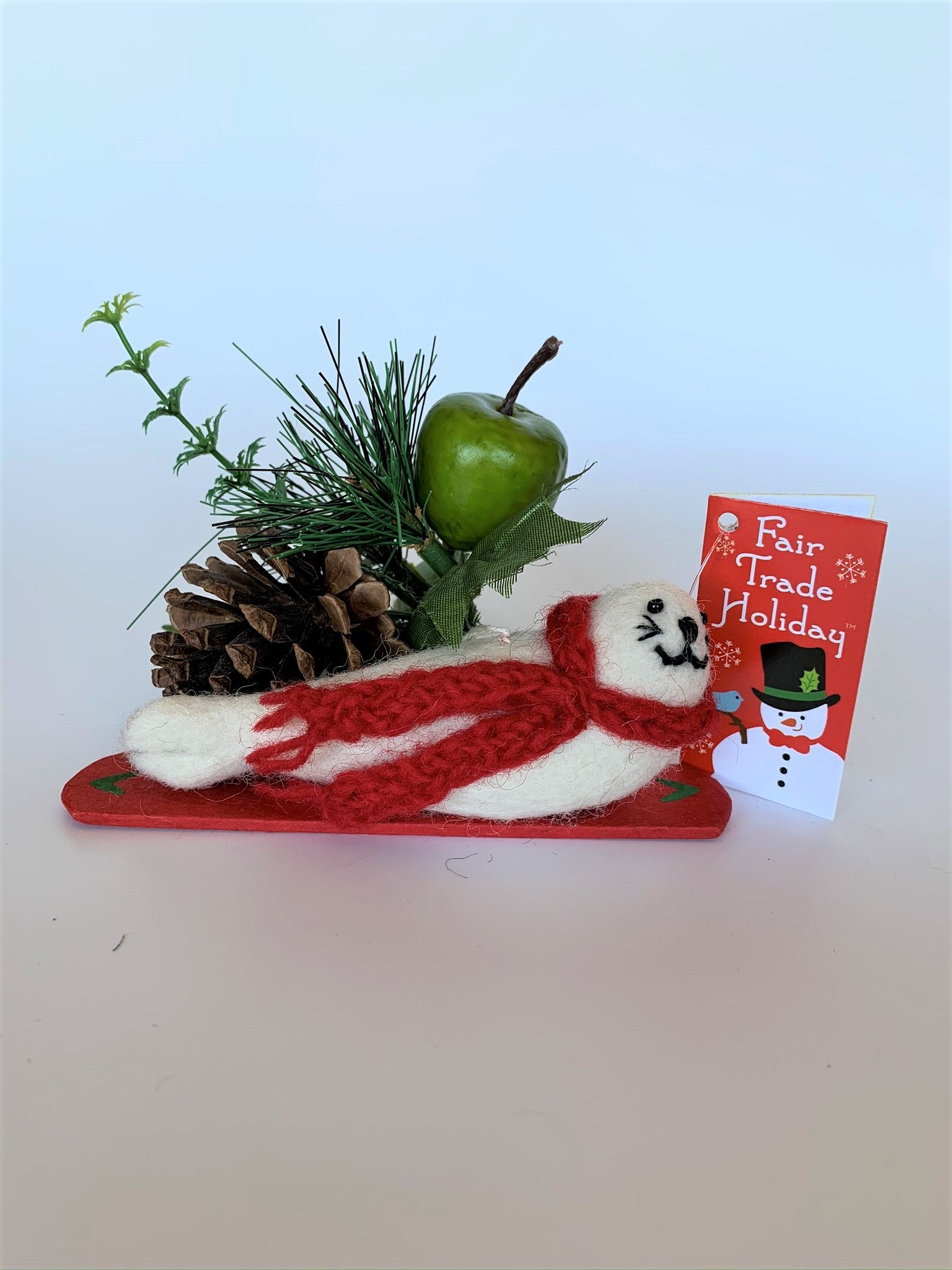 This is a side view of the snowboarding seal Christmas ornament that is handcrafted (fair trade) using 100% natural wool. The snowboard is handmade using paper that is hardened and feels almost like thin wood. It is off-white with black accents (eyes, nose, whiskers etc.), wears a red winter scarf and is lying on a red snowboard with green accents. Approximately 5.5"x2"x1.75". It comes with a fair trade holiday "to/from" tag to use if giving this as a gift.