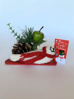 Load image into Gallery viewer, This is a side view of the snowboarding seal Christmas ornament that is handcrafted (fair trade) using 100% natural wool. The snowboard is handmade using paper that is hardened and feels almost like thin wood. It is off-white with black accents (eyes, nose, whiskers etc.), wears a red winter scarf and is lying on a red snowboard with green accents. Approximately 5.5&quot;x2&quot;x1.75&quot;. It comes with a fair trade holiday &quot;to/from&quot; tag to use if giving this as a gift.
