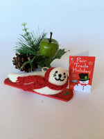 Load image into Gallery viewer, This is a snowboarding seal Christmas ornament that is handcrafted (fair trade) using 100% natural wool.  The snowboard is handmade using paper that is hardened and feels almost like thin wood.  It is off-white with black accents (eyes, nose, whiskers etc.), wears a red winter scarf and is lying on a red snowboard with green accents. Approximately 5.5&quot;x2&quot;x1.75&quot;.  It comes with a fair trade holiday &quot;to/from&quot; tag to use if giving this as a gift.
