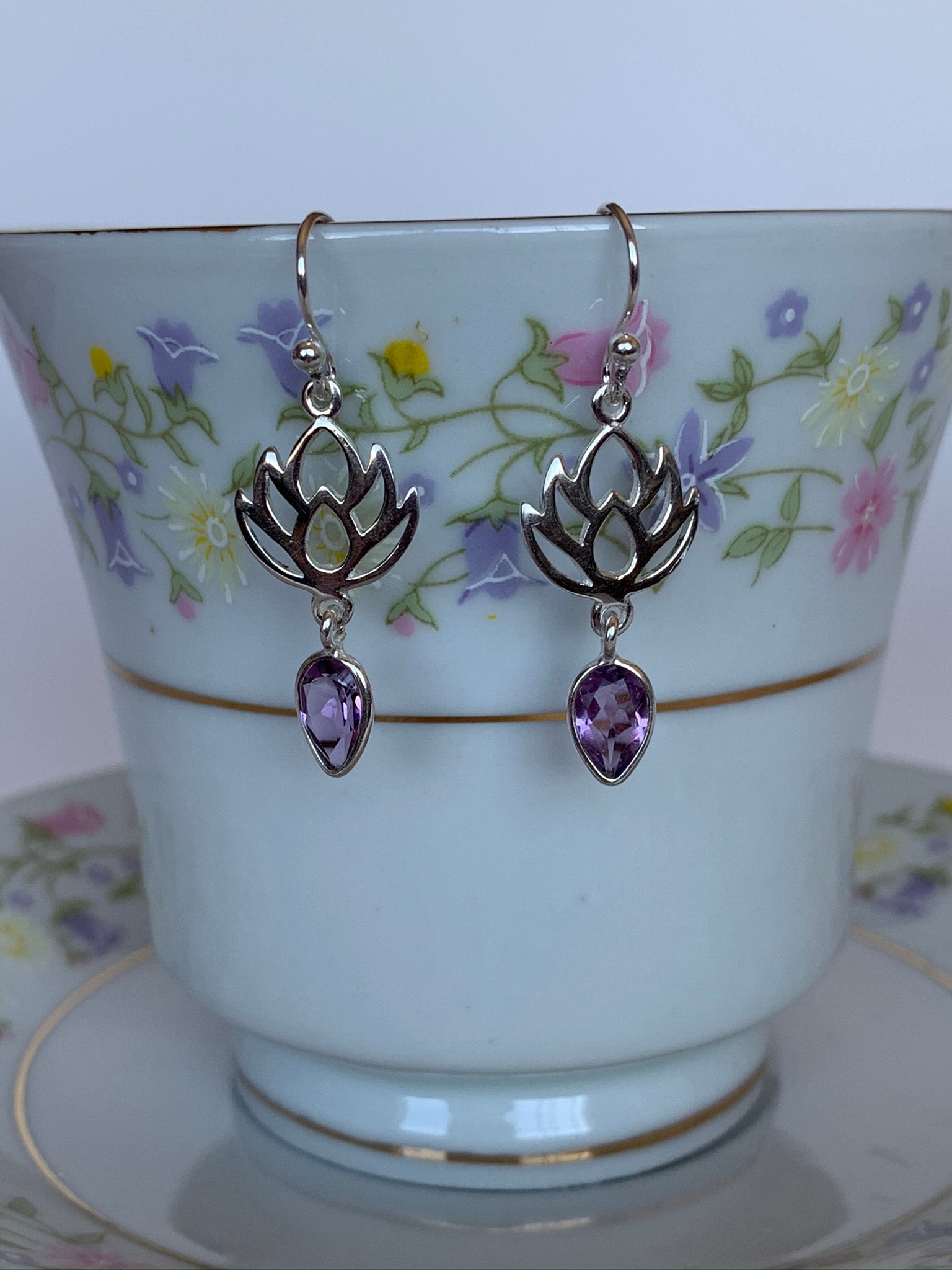 Close up of sterling silver earrings with earring wires, not posts. An amethyst (reverse teardrop) hangs from an open sterling silver lotus design. Earrings are lightweight and are approximately 1½" long.