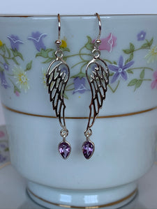 Close up view of lightweight sterling silver earrings. Small reverse teardrop amethyst gemstones hang from delicate silver angel wings. Wires, not posts, for wearing. Approximately 1¾" long.