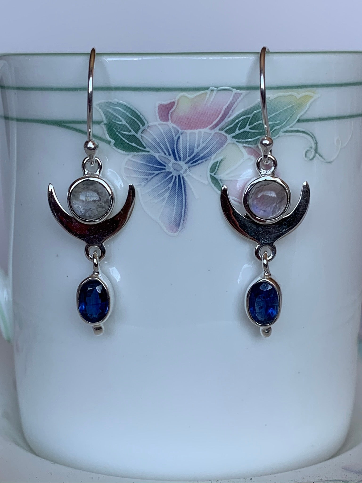 Close up view. These gorgeous & lightweight sterling silver earrings consist of a sterling upturned crescent moon with a small round moonstone within its curve and a blue oval faceted kyanite gemstone hanging below it. Wires, not posts for wearing. Approximately 1½" long.