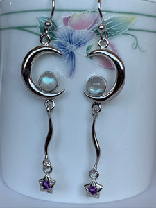 Close-up view of sterling silver Moon star earrings. Each earring displays a solid sterling crescent moon with a small, round moonstone in its lower curve and has a long, thin, curving sterling connector holding a small sterling star with a tiny amethyst at its center. Earrings are lightweight, have wires, not posts and are approximately 2" long.