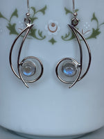 Load image into Gallery viewer, Close-up view. These sterling silver earrings have open (stylized) crescent moons with a sterling circle at the bottom, holding a small round moonstone. They are lightweight and approximately 1¾&quot; long.
