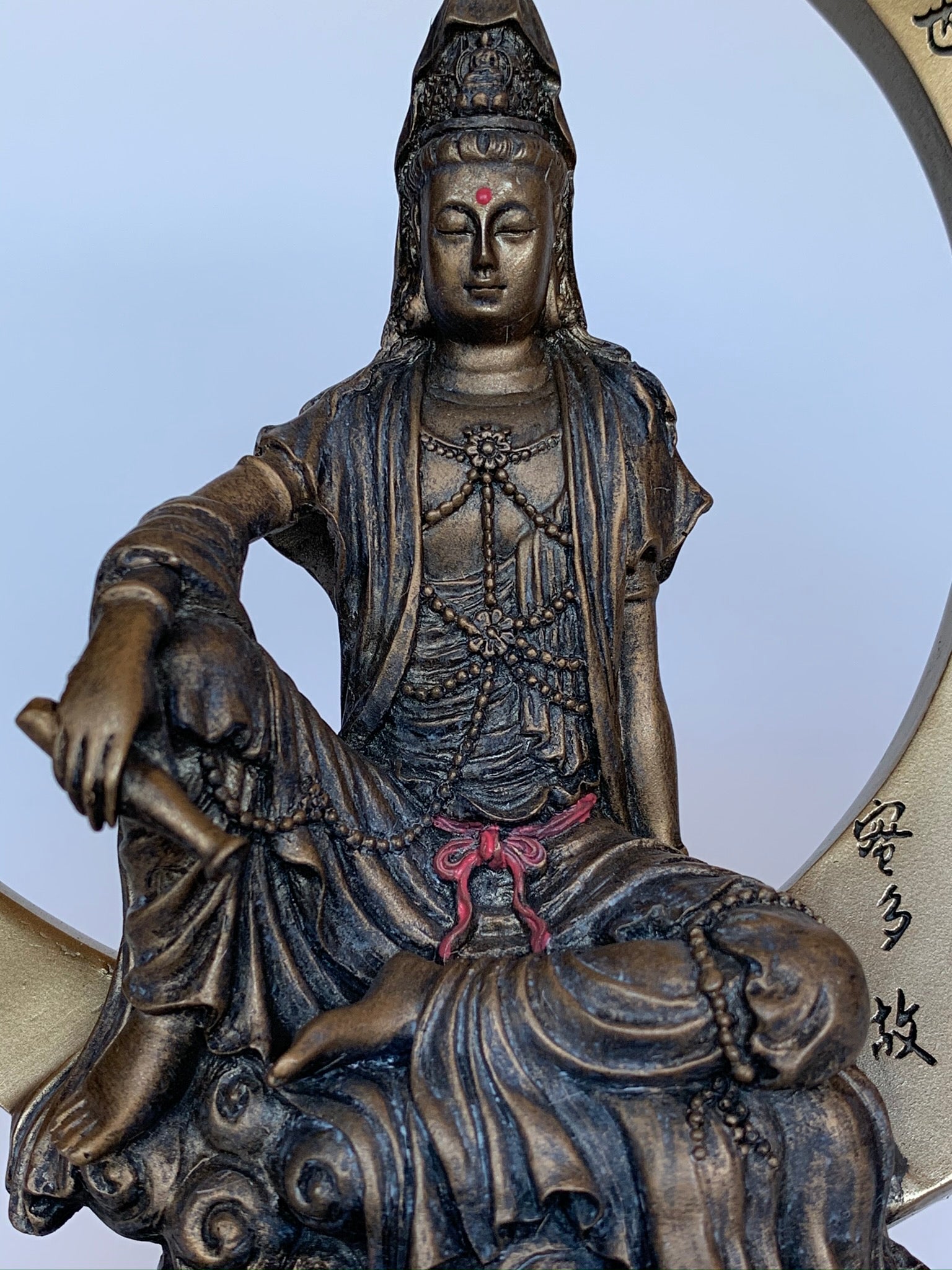 Close-up View of top of the beautiful, detailed statue of Kwan Yin sitting on a large crescent moon finished in a burnished bronze. Kwan Yin is a revered and powerful goddess. She symbolizes compassion, mercy and kindness.  The moon represents powerful feminine energy, intuition, emotions/feelings. Color is added to certain details on this lovely statue, adding to its appeal. Red and green are added to the lotus flower and leaves below her and red is added to her belt & forehead. Approximately 7¾".