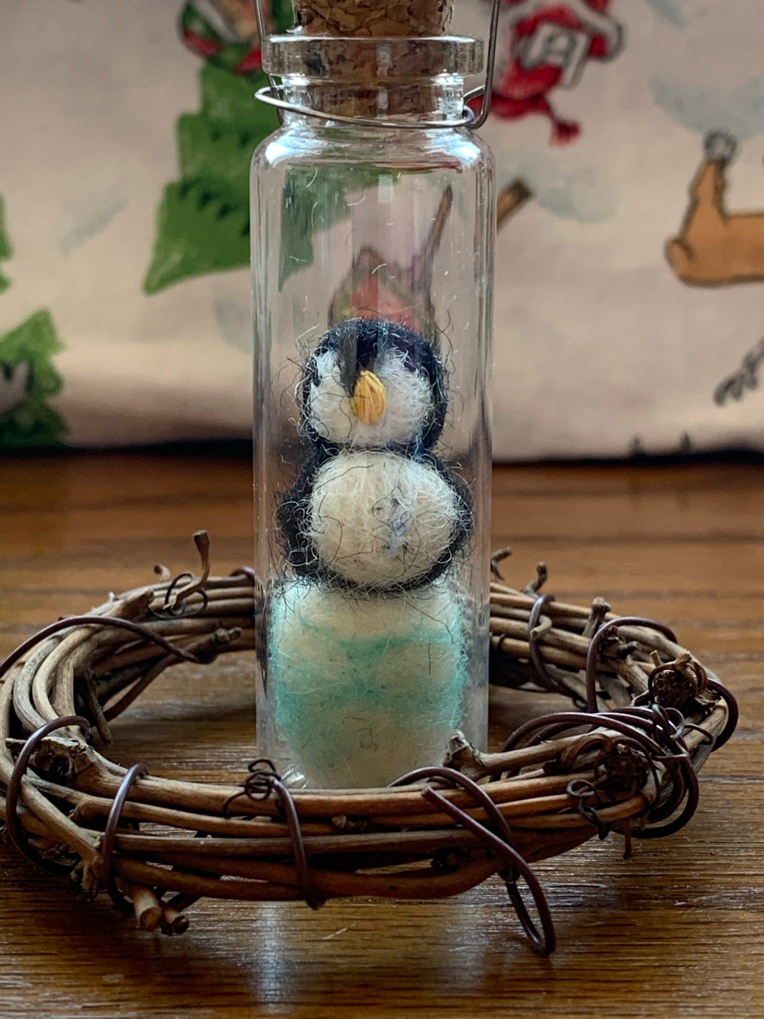 This is a close-up photo of the penguin bottle ornament that is handcrafted and the penguin inside the bottle is made of 100% natural hand-felted wool. The penguin is black and white and sits atop a blue and white iceberg. He is enclosed in a small, glass, corked apothecary bottle which is affixed with a metal wire to hang it on your tree , but you can also place it on a shelf or table top. Approximately 3"x1" (it is 6.75" if you include the wire hanger). It comes with a fair trade home and and garden tag.