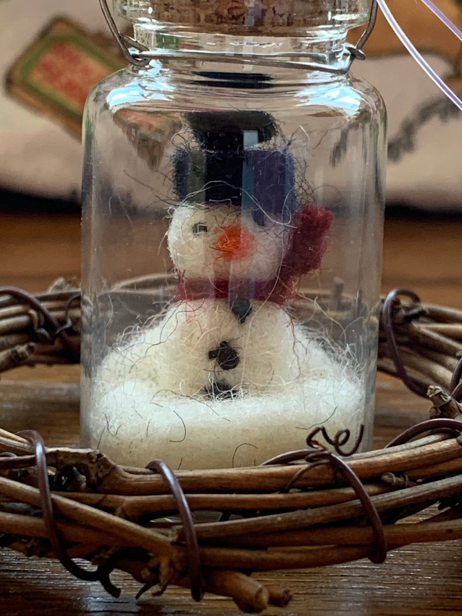 This is a close-up of the snowman bottle Christmas ornament. The snowman inside the bottle is handmade (fair trade) of 100% natural wool. He is off-white with a black hat and accents, an orange carrot nose, with a red winter scarf around his neck. He sits inside of a glass, corked apothecary bottle and has a wire hanger affixed for hanging as an ornament, but you can sit him on any shelf or table top as well. He comes with a fair trade holiday tag to use if giving as a gift.