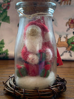Load image into Gallery viewer, This is a story jar Santa Christmas Ornament. A corked, apothecary jar holds Santa sitting on the snow holding a gift and with two other gifts on the snow around him. He wears the traditional Santa colors of red &amp; white and wears white woolen mittens. He is made of 100% natural, hand-felted wool and is approximately 4.5&quot;x2&quot;. He comes with a fair trade home &amp; garden tag.
