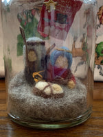 Load image into Gallery viewer, This close-up of the Nativity story jar Christmas ornament displays Mary, Joseph and baby Jesus with a star above them, sitting on the ground. They sit inside a corked apothecary bottle. The Nativity is made of 100% natural wool. The bottle is affixed with a metal wire for hanging, but can also be placed on a shelf or table top. Approximately 3&quot;x1&quot; (6.75&quot; if you include the wire hanger). It comes with a fair trade home and and garden tag.
