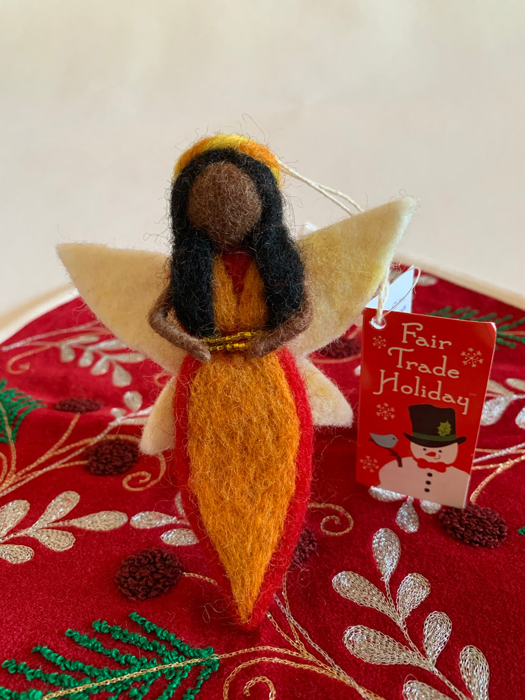 This fire-fairy Christmas ornament is handmade (fair trade) from 100% wool. She is black-skinned & wearing an orange dress with red on the edge & has a double row of tiny gold beads around the empire waist. She wears a yellow & orange headpiece, has black hair and white wings with a bit of light yellow on them. She has bendable arms and no facial markings. She is approximately 5"x4" and comes with a fair trade holiday "to/from" tag to use if giving as a gift.