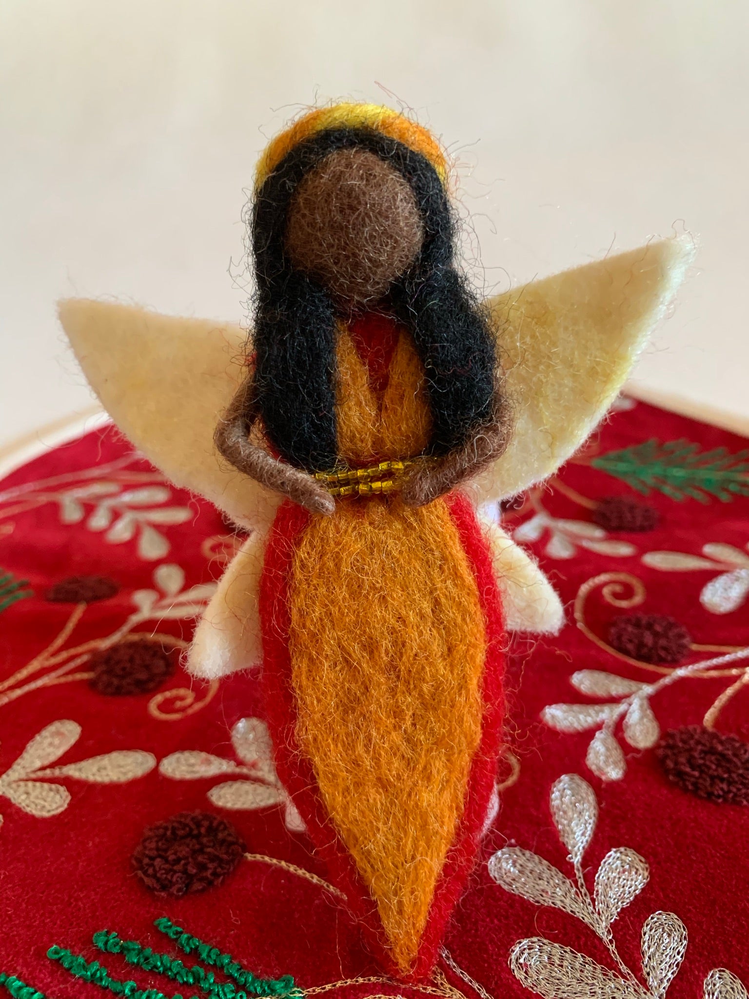 The close-up of the fire-fairy Christmas ornament is handmade (fair trade) from 100% wool. She is black-skinned & wearing an orange dress with red on the edge & has a double row of tiny gold beads around the empire waist. She wears a yellow & orange headpiece, has black hair and white wings with a bit of light yellow on them. She has bendable arms and no facial markings. She is approximately 5"x4" and comes with a fair trade holiday "to/from" tag to use if giving as a gift.