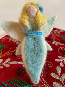 The close-up view of the air-fairy Christmas ornament is handmade (fair trade) from 100% wool. She is light/fair skinned & wearing a light blue dress with white on the edge with a double row of tiny blue beads around the empire waist. She wears a two-toned blue flower in her yellow hair & has white wings with a bit of light yellow on them. She has bendable arms and no facial markings. Approximately 5"x2.5" & comes with a fair trade holiday "to/from" tag to use if giving as a gift.