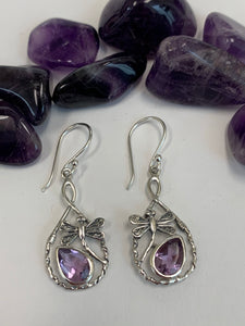Close-up view. Teardrop shaped amethyst set, at a slant, at the bottom of an open tear drop shaped silver loop. sterling silver dragonflies "fly" above the amethysts. These beautiful and fanciful earrings are lightweight, have wires, not posts and are approximately 1½" long