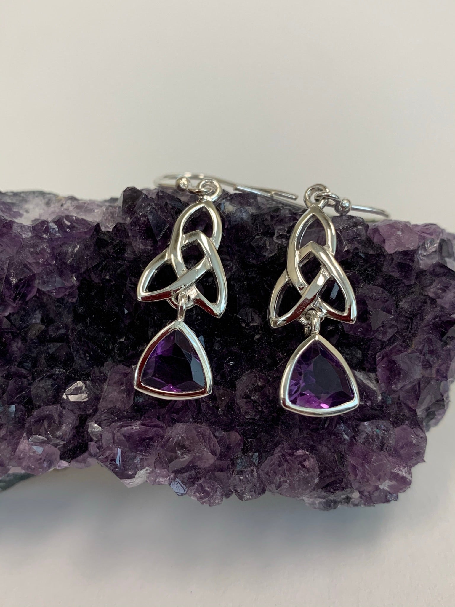 Trilliant cut (triangular) faceted amethyst gemstones outlined in sterling silver, dangle from an elongated Celtic trinity knot, or triquetra. These earrings are lightweight, have wires, not posts and are approximately 1½" long.