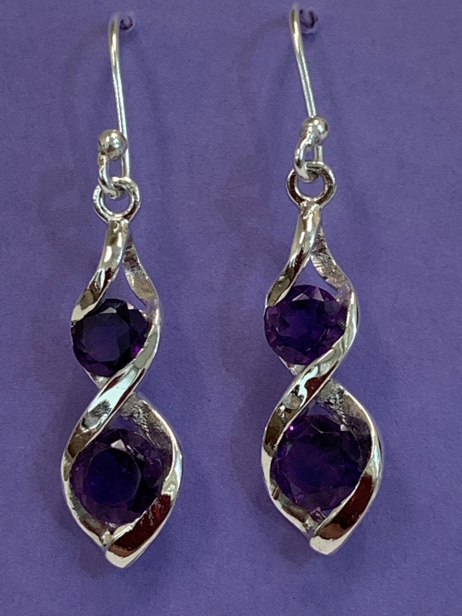Close-up view. Two amethyst gemstones are set into each of the sterling silver earrings in a "twister" setting (like a stylized figure 8), one in the top part of the eight and one in the bottom. These earrings are lightweight, have wires, not posts and are approximately 1¼" long.